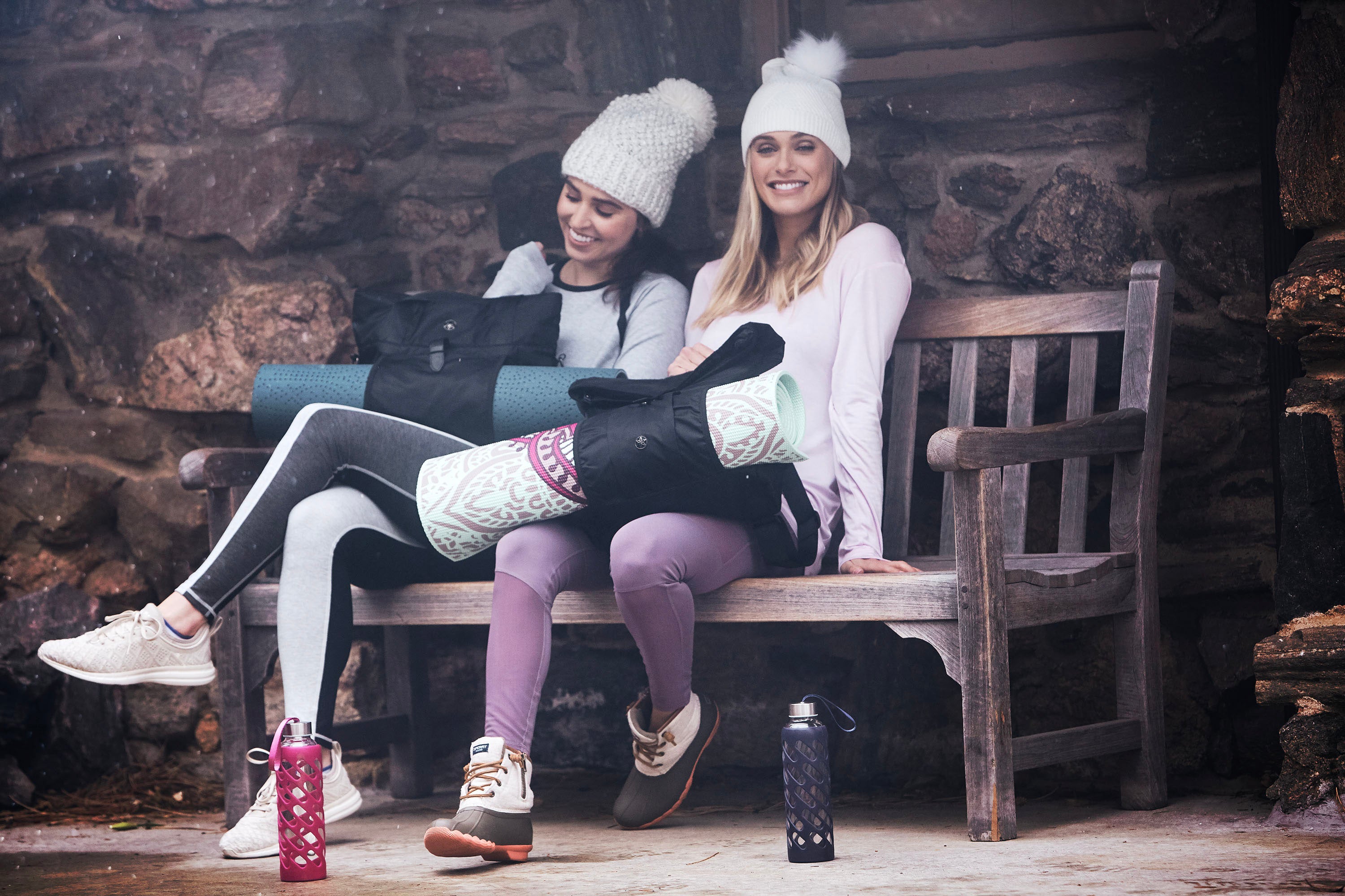 A blonde and brunette sit wearing winter caps and yoga clothes on a bench outside a stone building in a wintery setting with bags holding yoga mats on their laps