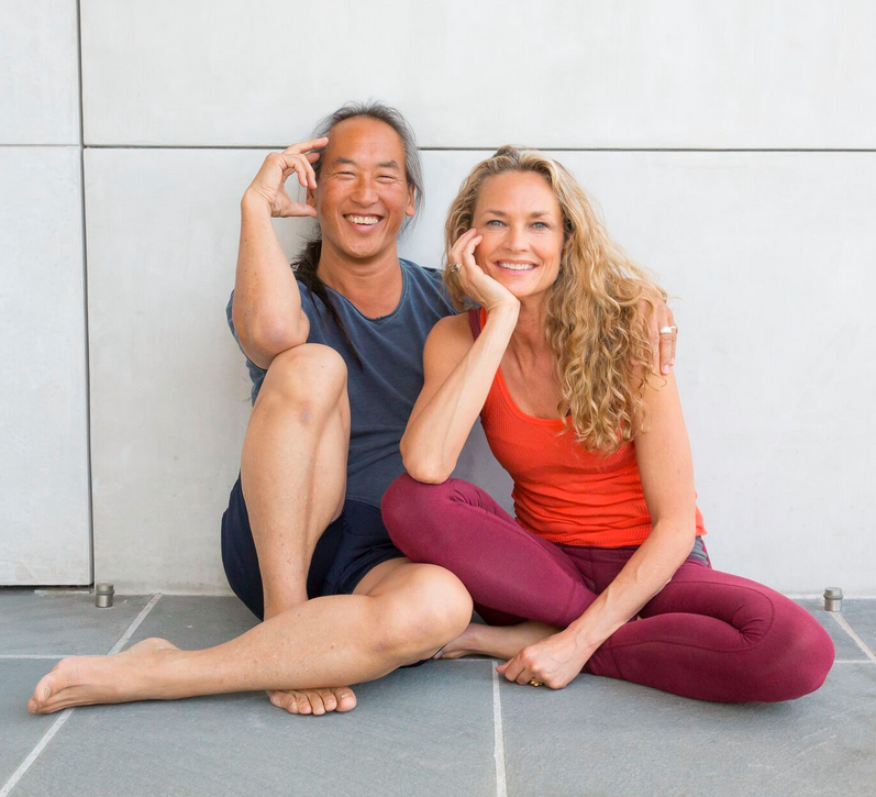 Middle aged couple sit cross-legged on a grey tile floor against a white wall in yoga clothes