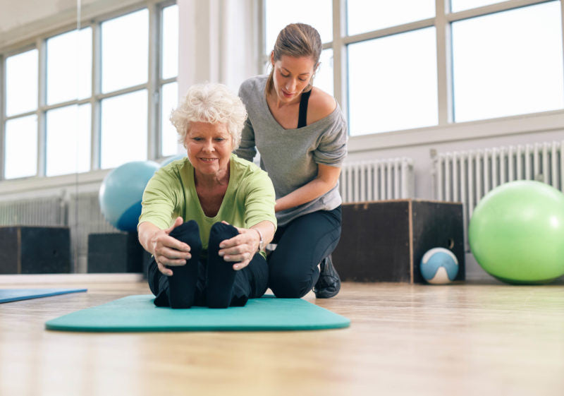 Yoga teacher adjusts elderly woman in seated forward fold