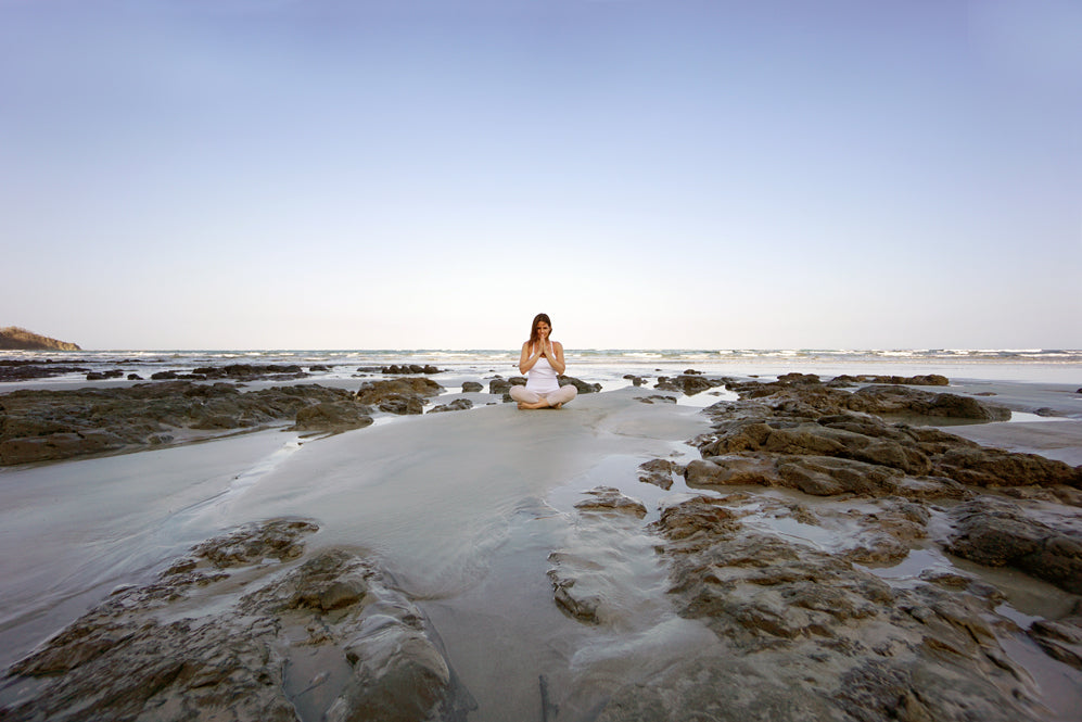 woman with dark hair meditates on the beach in white yoga clothes