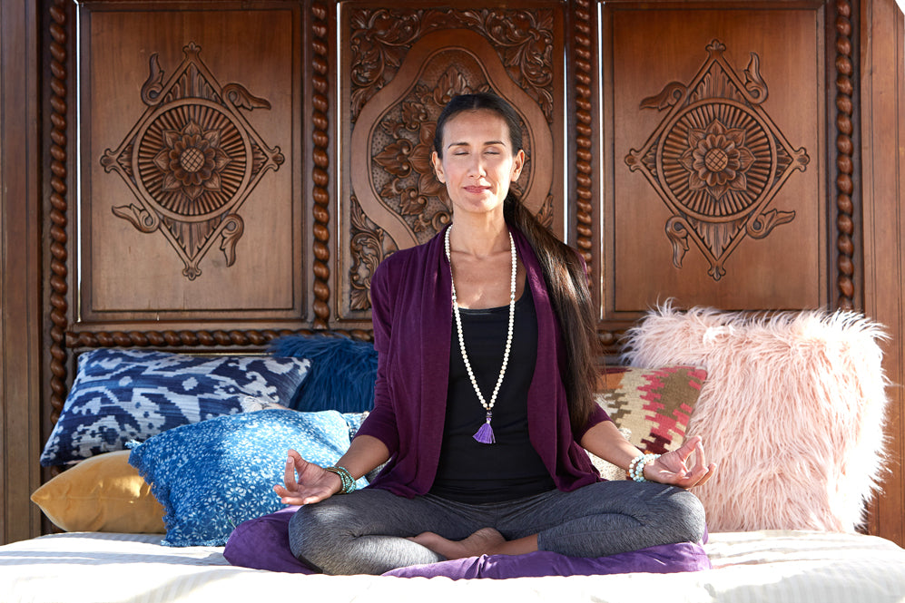 Woman sits and meditates on a bed wearing malas