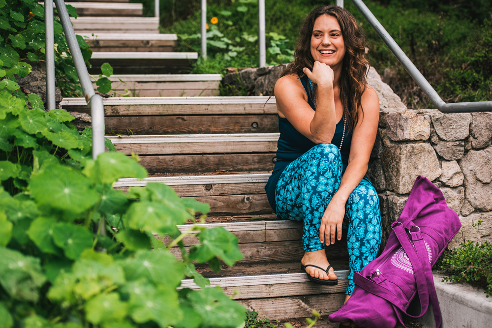 woman sits smiling with yoga mat on outdoor stairs